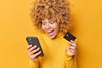 Smiling surprised curly haired young woman stares at smartphone holds credit card does online...
