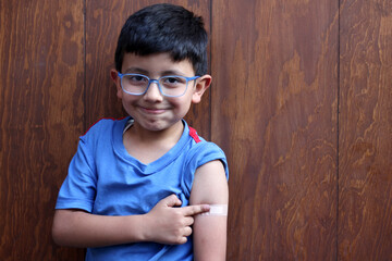 Little 6-year-old Latino boy with glasses and a blue shirt shows his arm with a bandage because he...