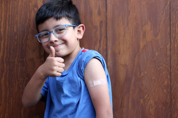 Little 6-year-old Latino boy with glasses and a blue shirt shows his arm with a bandage because he...