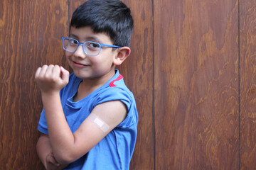 Little 6-year-old Latino boy with glasses and a blue shirt shows his arm with a bandage because he...