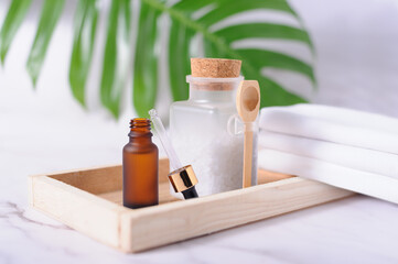 Plastic bottle filled with white bath sea salt and wooden spoon, amber color dropper and white towels on wooden tray.