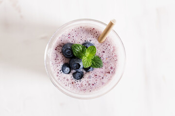 Glass of blueberry milkshake with fresh blueberries on white wooden background.