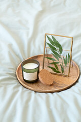 Green scented candle, golden photo frame with green leaves and wooden tray stand on bed with green linen.