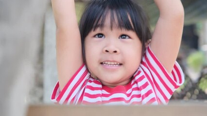 Cute Asian little girl hangs a wooden bar with her hands for exercise in the backyard playground. Active child hangs on bar with two hands.