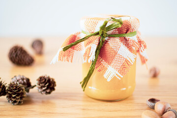 Natural fabric decorating a jar with honey. Bright warm pastel colors, pine cones on a wooden table. Selective focus on the details, blurred background.