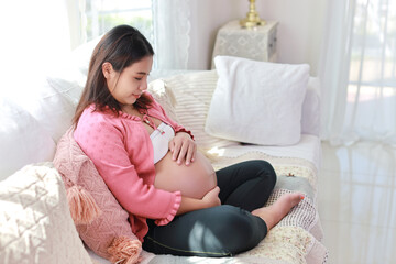 Happy smiling young asian pregnant woman sitting and resting on sofa in living room while touching and looking her belly. Expectant mother preparing and waiting for baby birth during pregnancy.