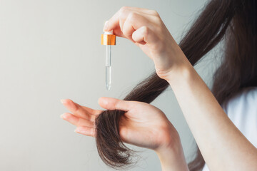 A woman with dark hair applies a cosmetic product to the ends of her hair with a pipette. Close-up of the hands. Gray background. The concept of hair care