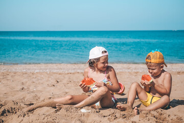 Portrait of children of sister and brother on the beach with watermelon. Girl and little boy are eating on the beach. The children had a picnic by the sea. Copy space.