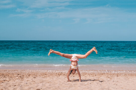 The girl makes a gymnastic wheel element on the background of the sea. The child is resting on the beach in summer. Summer time. Travel.