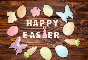 Easter greeting card with pastel coloured homemade cookies shaped in bunnies, butterflies, eggs, carrots and flowers. Top view of traditional Easter signs and symbols on dark brown.