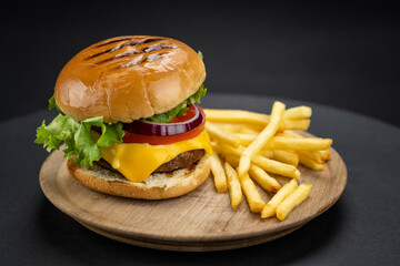 Tasty homemade beef burger with fresh ingredients served on little wooden cutting board with french fries on dark background, close-up view..