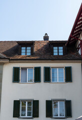 Regular white old tenement house with small attic window on the brown tiled rooftop and dark green wooden shutters. Residential building seen on a sunny summer day.