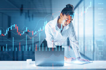 Attractive caucasian female working at desktop with laptop and glowing business chart on blurry office interior background. Trade, finance, market and financial growth concept. Double exposure.