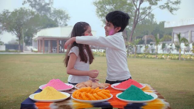 Cute Indian boy and a little girl applying powder color on each other's cheeks - organic Gulal  a spring festival. Adorable siblings embracing each other outdoors while wishing a Happy Holi - famil...