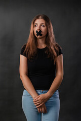 Portrait of a young woman with her mouth sealed with black tape, restriction of freedom of speech, censorship and a ban on speaking and expressing her opinion, isolated on a dark background. - obrazy, fototapety, plakaty