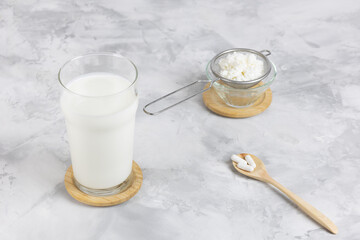 A glass of kefir and kefir grains on a strainer over a glass jar, standing on a wooden cutting...