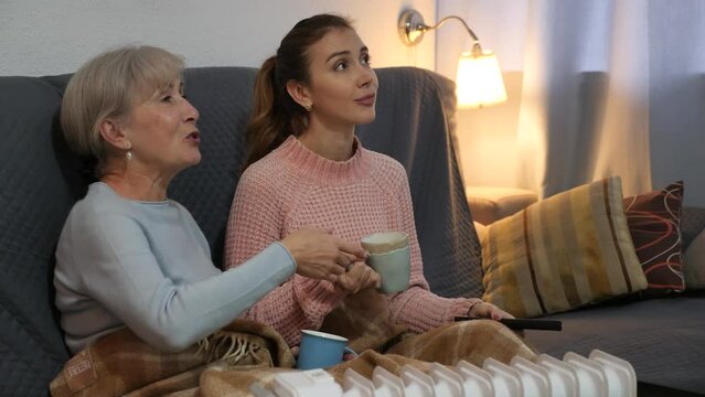 Home cozy evening. Positive young girl relaxing with aged mother, sitting on sofa in plaid blanket with cups on tea and watching TV