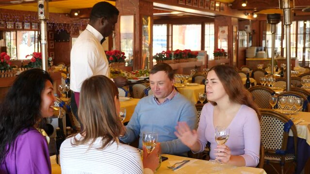 Group of people sitting at table in restaurant and waiting for food. African-american man waiter serving table.