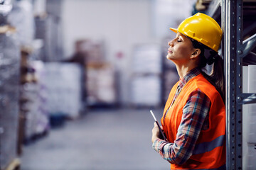 Tired female worker with tablet in hands leaning on shelf and taking a break in warehouse.