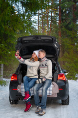 A young couple taking selfies with a phone and enjoying beautiful winter weather in the open trunk of a car in the winter snowy forest. Happy caucasian family enjoying the romantic outdoor date.