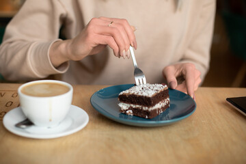 Fototapeta na wymiar Cropped photo of woman hands in coffee shop cafe, eating tasty brownie powdered sugar. Cup of coffee on wooden table. 