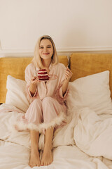 Obraz na płótnie Canvas Full photo of joyful young european woman in pajamas looking at camera while eating in bed alone. Girl blonde cute smiling holding plate with cake in her hands. Unhealthy but tasty food concept.