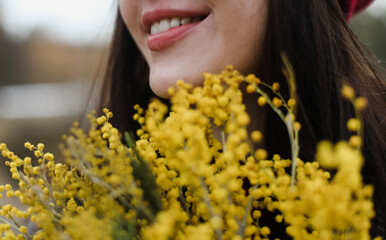 close-up portrait of a smiling brunette girl with red lips enjoys a large bouquet of fresh spring yellow mimosa bouquet outdoors