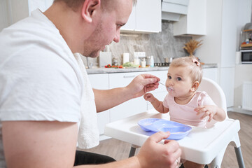 Fototapeta premium Baby girl in pink clothes during the feeding from the spoon in the kitchen