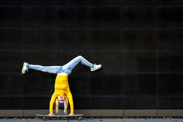 Athetic young woman upside down on a skateboard, black wall background, yellow shirt and blue...