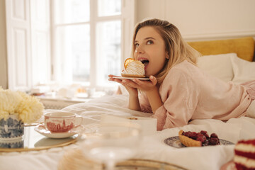 Lovely young caucasian girl appetizingly bites cake, looking up, lying in bed on her stomach. Beauty blonde woman in light pink pajamas is enjoying dessert. Sweets lover concept