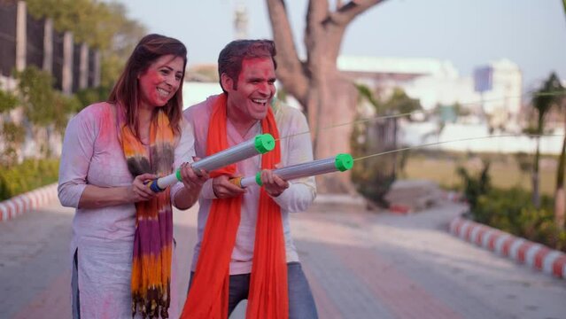 A young couple playing with a water gun during Holi celebrations - Pichkari  water splash  childhood games. Indian couple in an ethnic dress and with Gulal color on their faces - best friends  a pu...