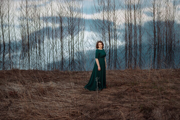 Woman in a long dress stands by the water, river, lake, which reflects bare trees, early spring or autumn © Antonina Trushina
