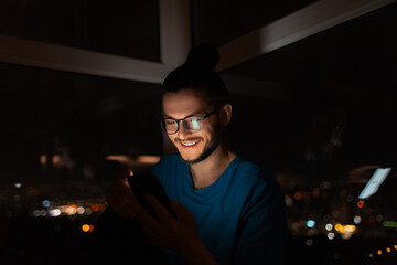 Night portrait of smiling man looking in smartphone on background of window.