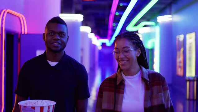 Happy young african american woman and man walking by neon hall with bucket of popcorn, chatting at cinema