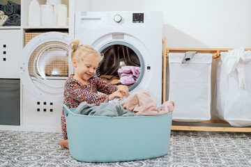Cute little girl with blonde hair, small child sits in bathroom, laundry room by large bowl full of clothes, daughter throws things into washing machine, helps mom with daily chores © ABCreative