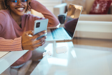 Cheerful african student black woman sitting at table studying using laptop reading a book, take a break holding mobile phone surfing internet received message from friend chatting about weekend plans