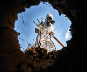 View from inside pit of ecologist digging pit by shovel and planting tree, wearing gas mask and...