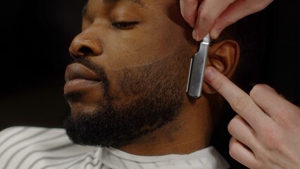 Man shaving beard of african american man with straight razor in barbershop