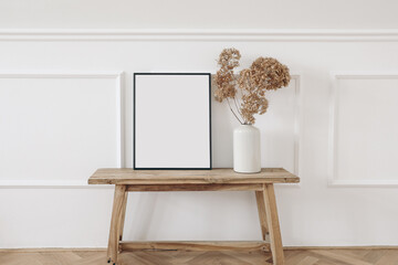 Vase with dry hydrangea flowers on old wooden bench. Blank black picture frame mockup. White wall moulding background, stucco decor. Empty copy space. Elegant interior. Summer, fall still life photo.