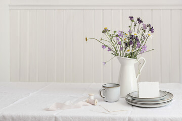 Spring, summer breakfast still life. Jug with wild flowers bouquet on linen table cloth. Daisies, bluebells, aquilegia. Greeting card mockup. Cup of coffee, ceramic plates. White wooden wall backround