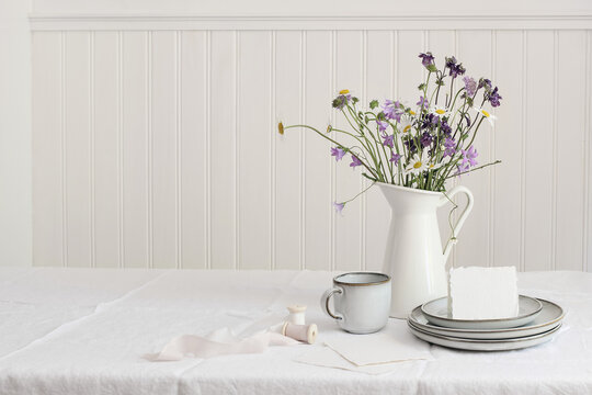 Spring, summer breakfast still life. Jug with wild flowers bouquet on linen table cloth. Daisies, bluebells, aquilegia. Greeting card mockup. Cup of coffee, ceramic plates. White wooden wall backround