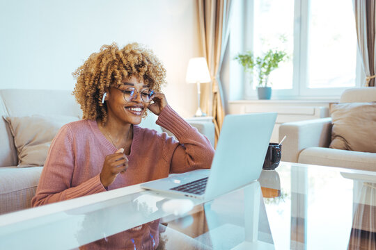 Smiling African American Woman Wearing Glasses and Wireless Earphones Makes a Video Call on her Laptop Computer at her Home Office. Smiling businesswoman in a video call using laptop in the office