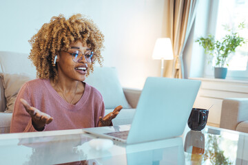Young woman video calling using a laptop sitting on a sofa wearing earphones. Using laptop and...