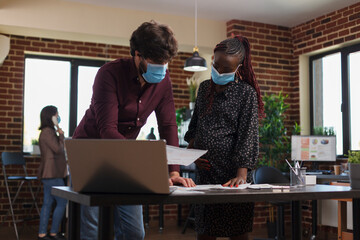 Pregnant businesswoman wearing facemask against covid reviewing project member list. Financial agency office colleague wearing protective mask and analyzing marketing strategy ideas for startup.