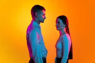 Portrait of two young people, man and woman in white cloth calmly standing, posing isolated over orange background in blue neon