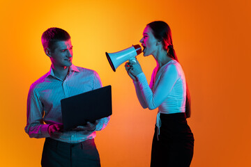 Portrait of young man, employee with laptop and woman shouting in megaphone isolated over orange background in neon lights