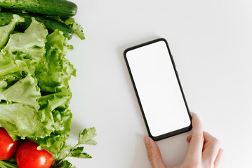 Mock up smartphone and eco vegetables, top view. Hand holding mobile phone with blank white screen and garden organic vegetables on white table. Healthy organic food concept with advertising space