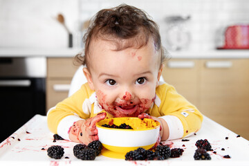 Happy baby with messy face eating blackberries in high chair
