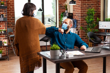 Businessteam with medical protective face mask to prevent infection with coronavirus touching elbow while working in startup office. Businessman analyzing data chart typing company strategy