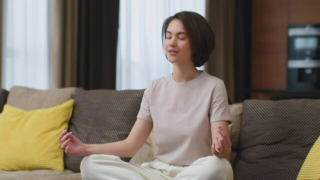 Peaceful young woman calming, breathing deep, meditating with mudra om gestures, doing yogs exercise sitting on sofa